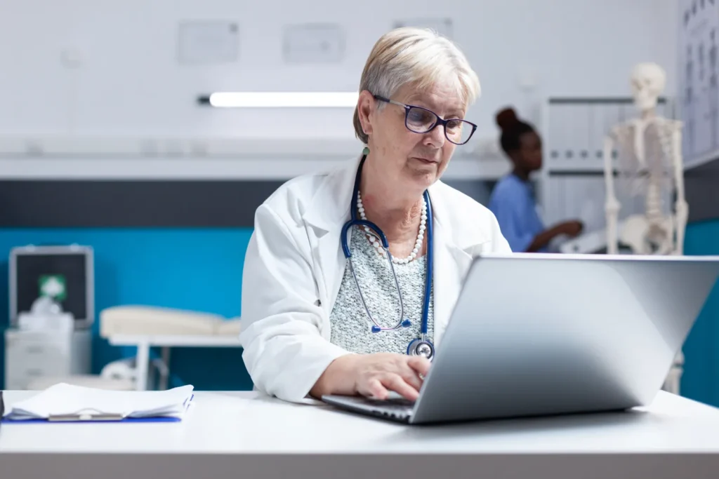 Senior physician working at a laptop in a clinic exam room