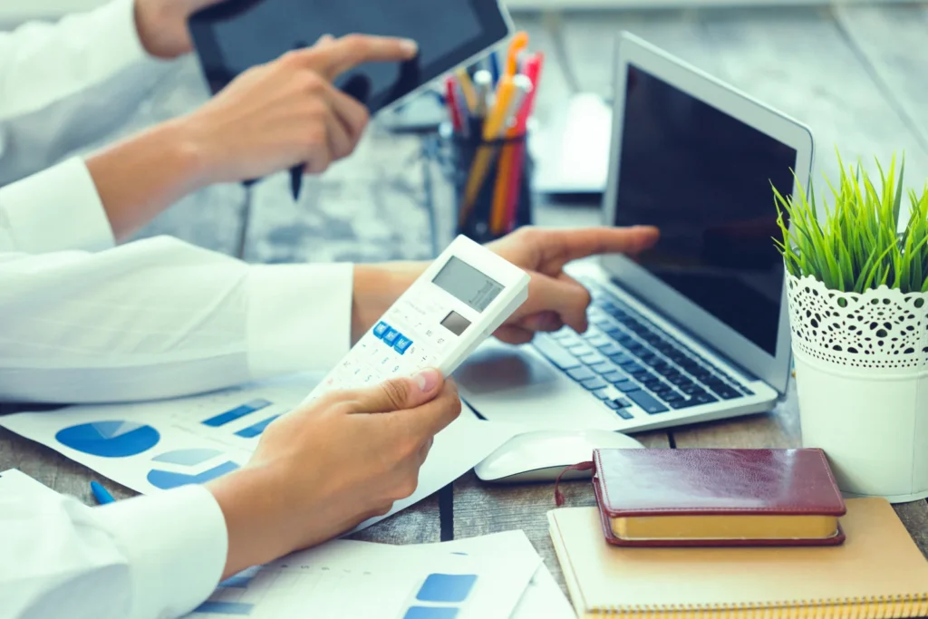 Two billing staff comparing payer reports on a calculator and a laptop