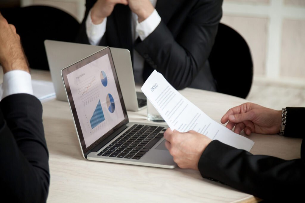 Hands passing a document across a table beside a laptop showing charts