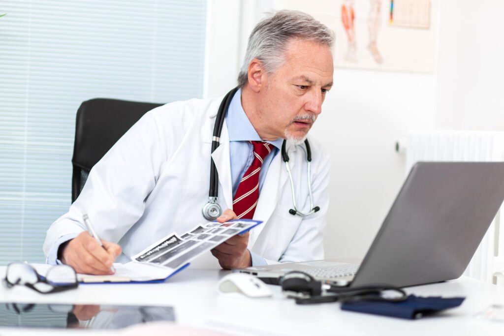 A senior physician reviewing patient records at a desk with a laptop and stethoscope