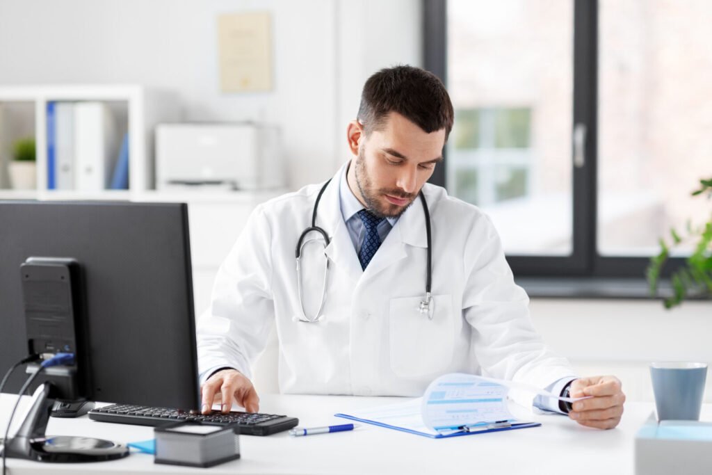A physician working at a desktop computer with documents and a calculator