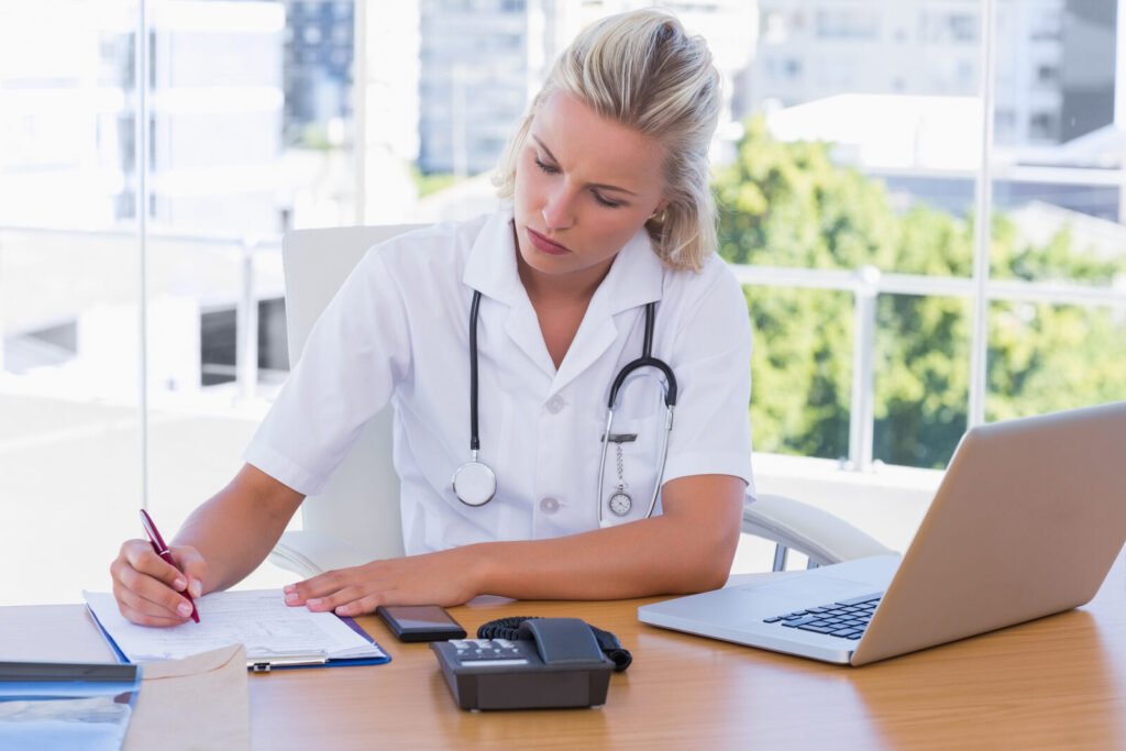 A clinician writing on paperwork beside a laptop by a window