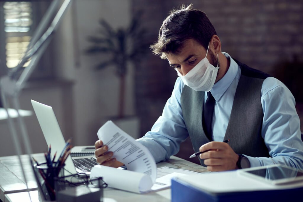 A masked billing specialist reading a printout at a desk lit by a monitor
