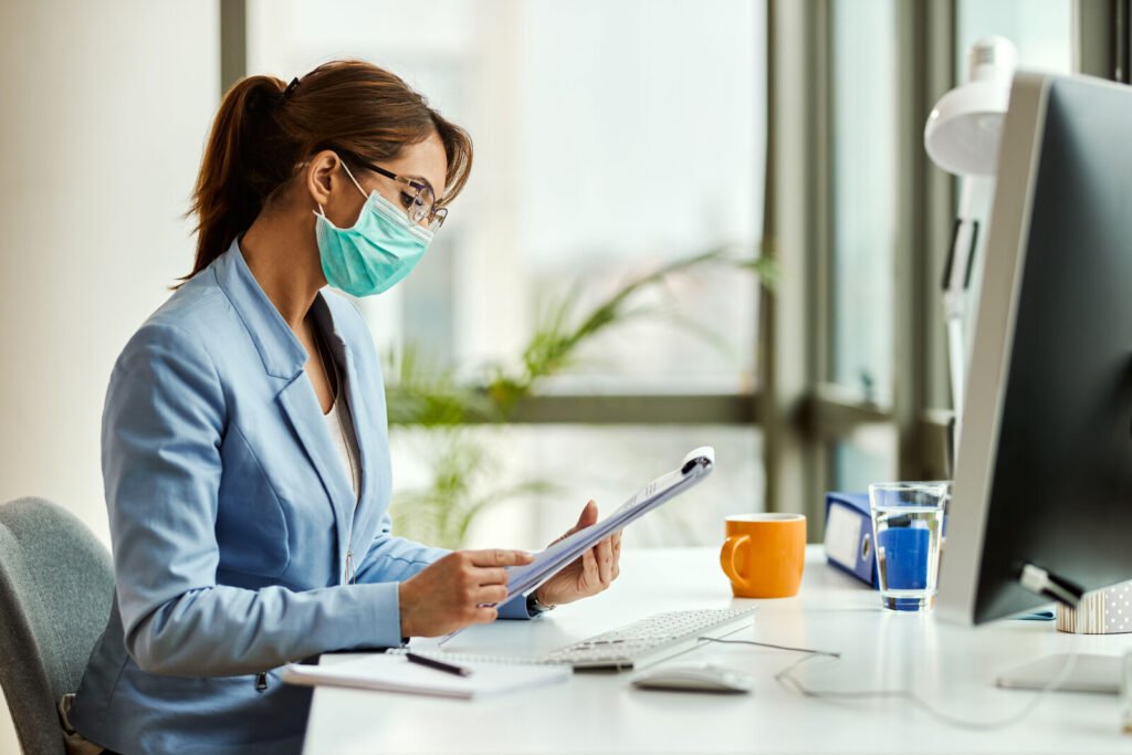 A masked billing specialist reviewing records on a clipboard at her desk