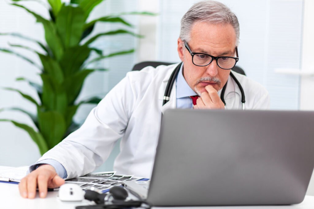 A physician in glasses thinking while looking at a laptop