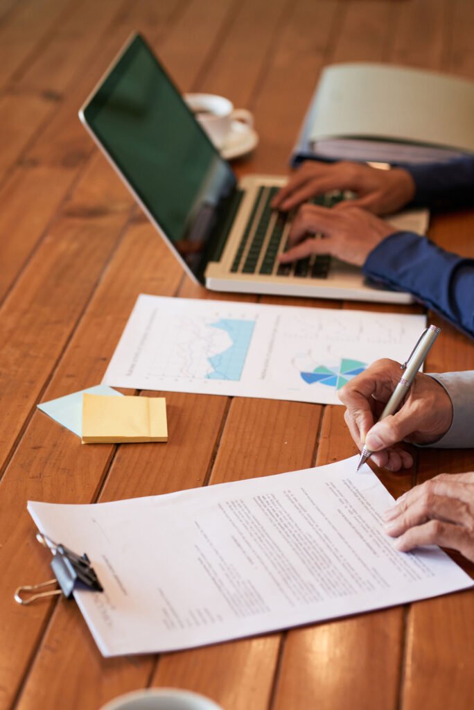 Hands signing compliance paperwork on a clipboard beside a laptop