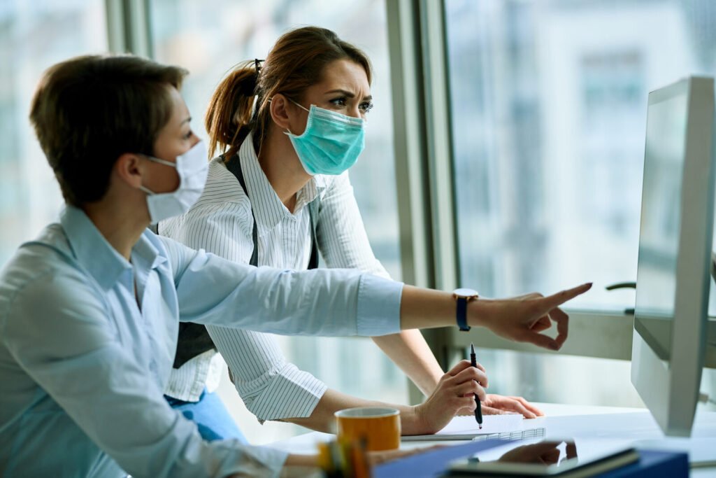 Two masked colleagues discussing information on a monitor at a shared desk
