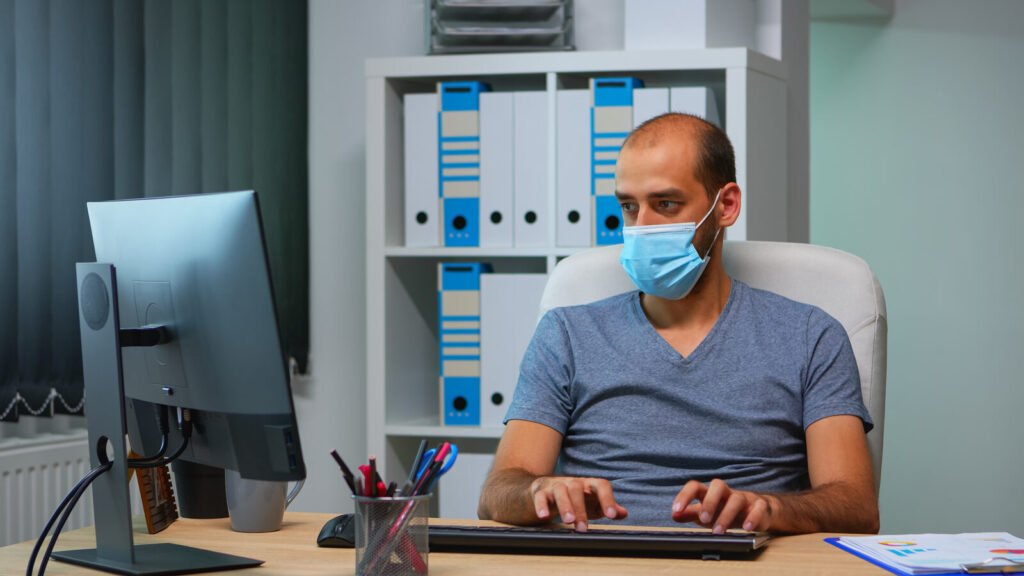 A masked specialist working at a desktop computer in an office lined with binders