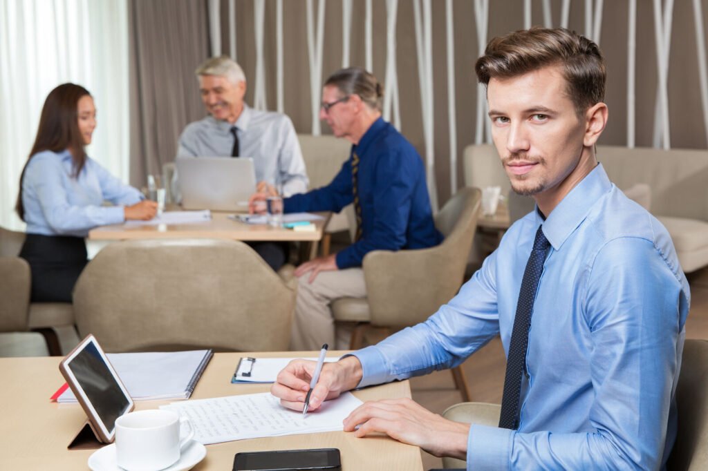 An administrator writing notes at a table while colleagues meet behind him
