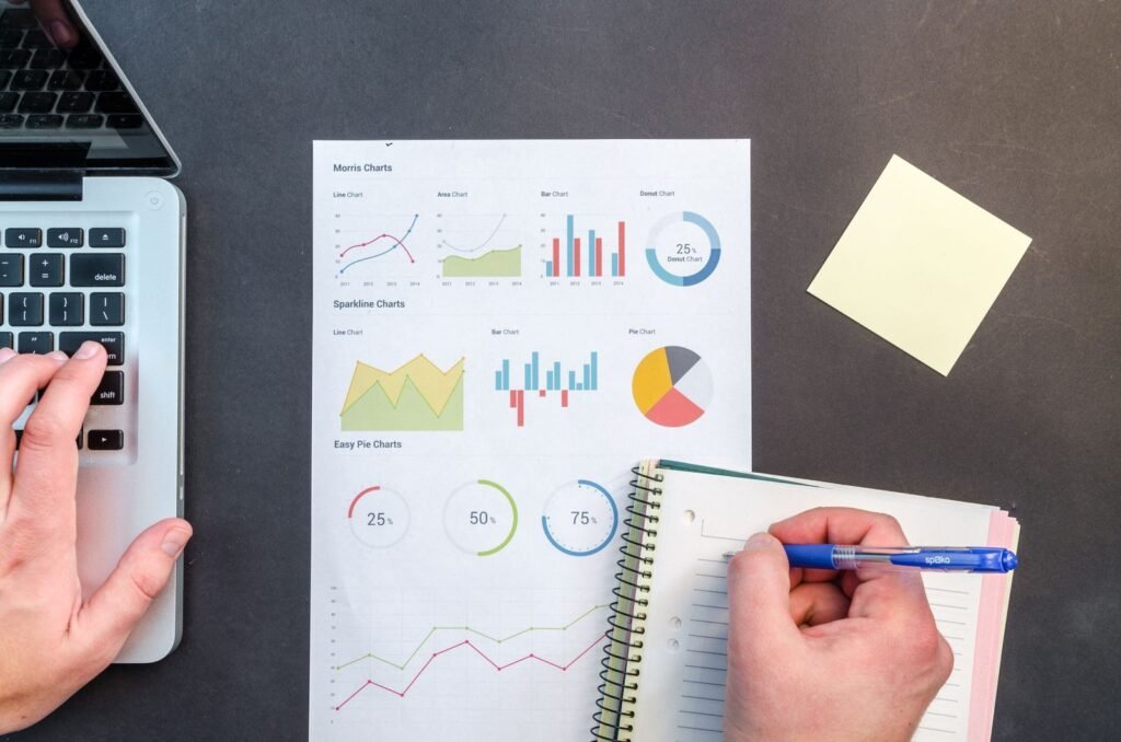 Hands writing notes beside printed billing charts and a laptop
