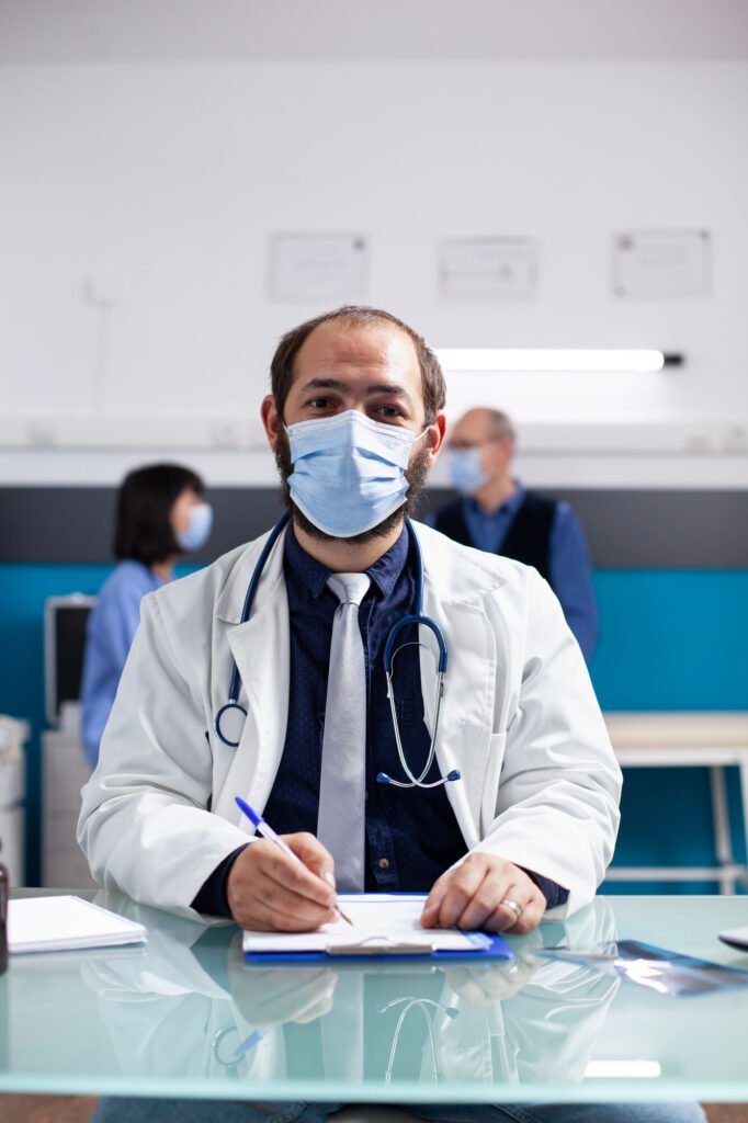 A masked physician writing at a glass desk in a clinic